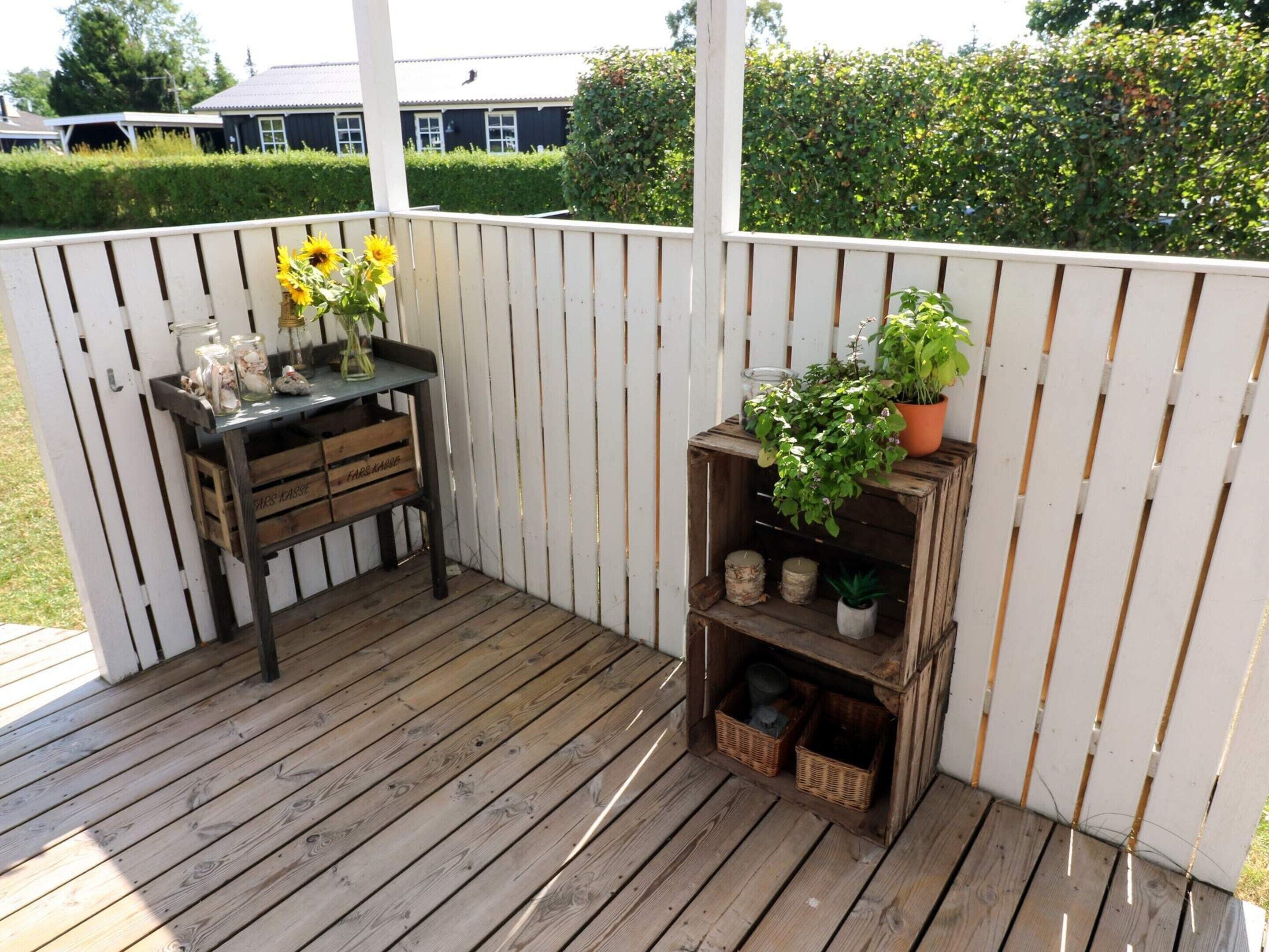 A wooden deck with a white fence, a small table with jars and a vase of flowers, and a shelf with plants and baskets.