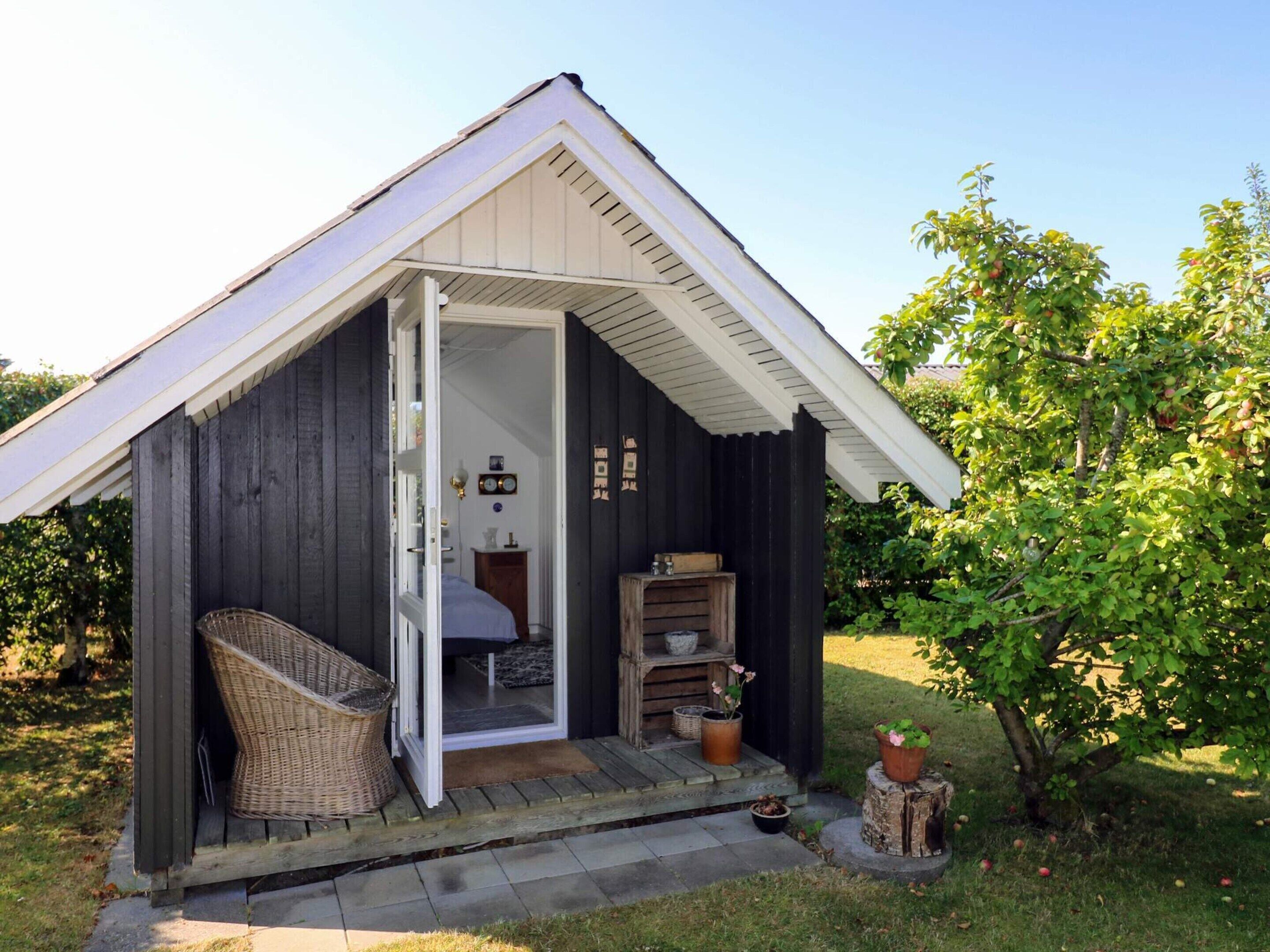 A small wooden cabin with a porch, a wicker chair, a bed, and a potted plant.