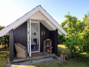 A small wooden cabin with a porch, a wicker chair, a bed, and a potted plant. - 8 Person Holiday Home in Hadsund (Hadsund)