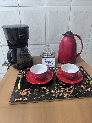 A coffee station with a black coffee maker, a red coffee pot, a glass container with coffee grounds, and two red cups with saucers.
