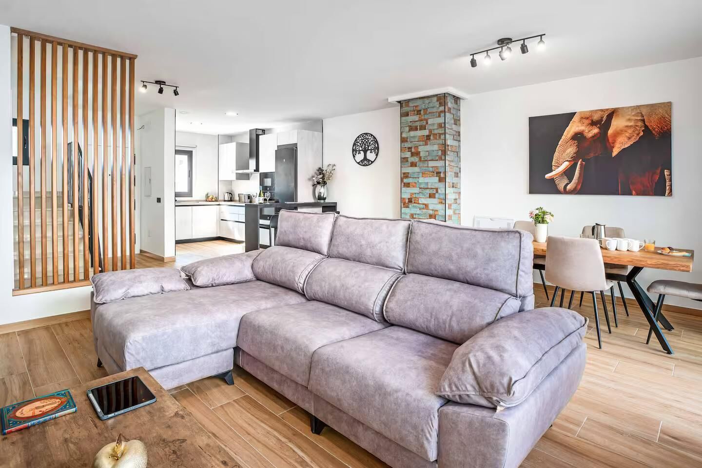 A modern living room with a grey sectional sofa, a dining table with chairs, a kitchen in the background, and a decorative wall piece.