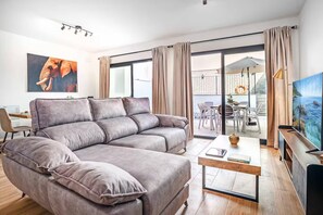 Una sala de estar moderna con un sofá gris, una mesa de centro y una televisión. Hay un balcón con mobiliario exterior y vistas al mar.