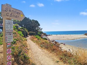 A wooden signpost with text and symbols indicating a beach area, a dirt path leading to the beach, and the ocean in the background. - Cozy studio for 2 people — Les Villas de la Plage, Rospico (Névez) (Névez)