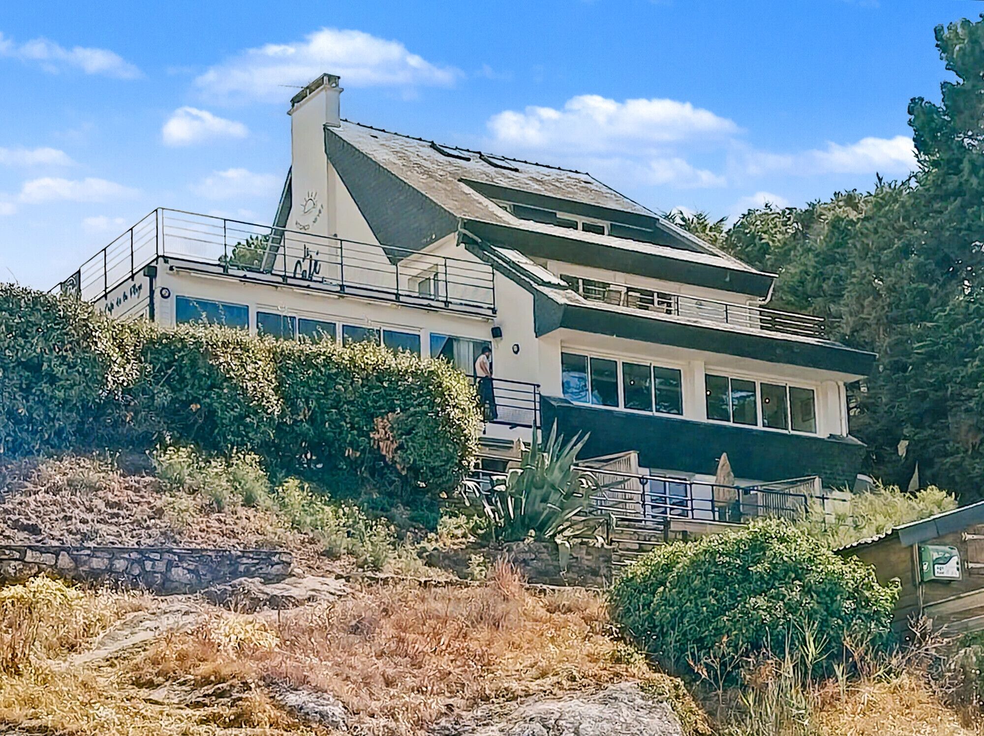 A two-story house with a balcony, surrounded by vegetation and a stone wall.