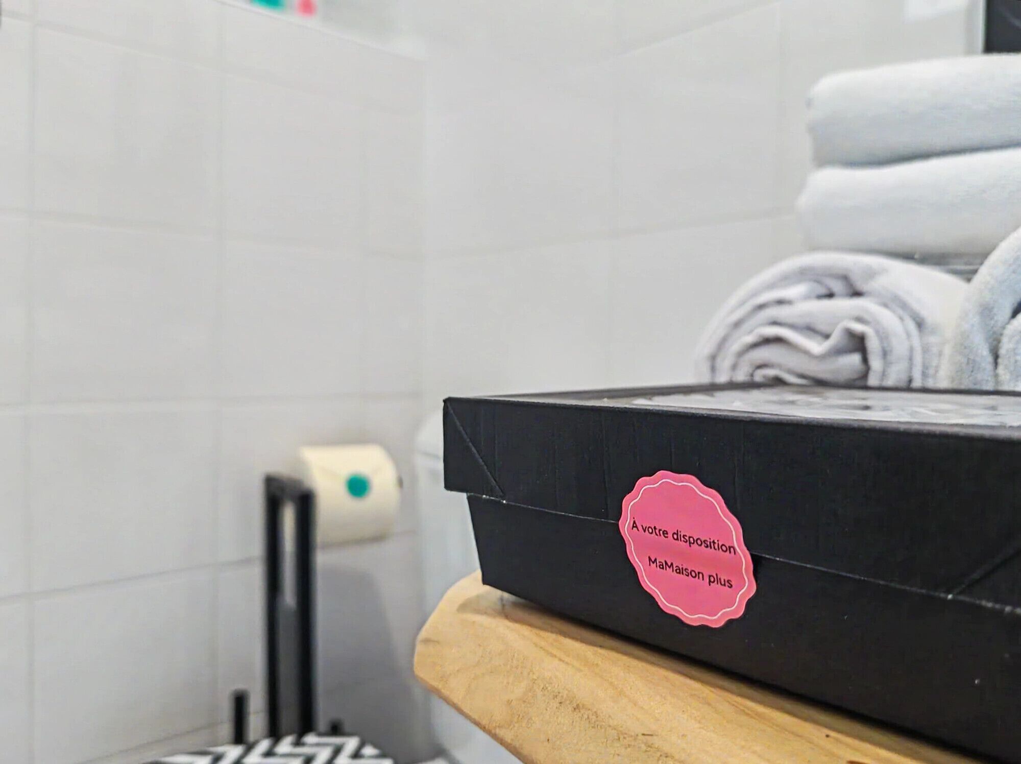 A bathroom with a black box on a wooden surface, a roll of toilet paper, and stacked towels.
