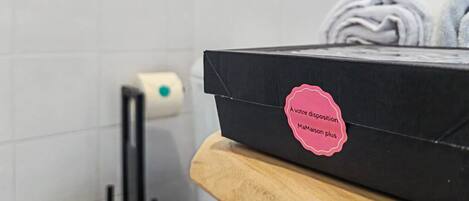 A bathroom with a black box on a wooden surface, a roll of toilet paper, and stacked towels.