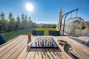 A chessboard on a wooden deck with a view of a grassy area and trees, a hanging chair, and a table with a mug and cookies. - Villa Naya - Three Bedroom Villa, Sleeps 6 (Geriş Mahallesi)