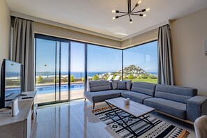 A modern living room with a large window, a sectional sofa, a flat-screen TV, and a pool view. - Villa Nerea - Three Bedroom Villa, Sleeps 6 (Geriş Mahallesi)