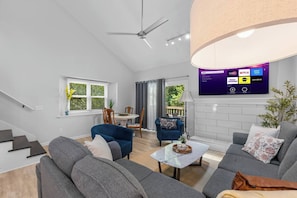 Ein modernes Wohnzimmer mit Wand-Fernseher, der Streaming-Dienste anzeigt, Deckenventilator und Essbereich mit Tisch und Stühlen.