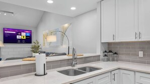 A modern kitchen with a built-in dishwasher, white cabinetry, a large flat-screen TV displaying streaming services, and a map on the wall. - Coastal Gem near Wrightsville Beach, Golf & Dining (Wilmington)