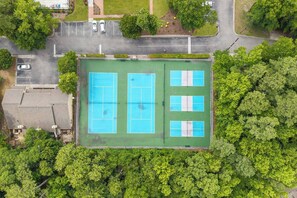 Luftaufnahme eines Sportkomplexes mit mehreren Tennisplätzen, umgeben von dichter Vegetation und angrenzenden Parkflächen.