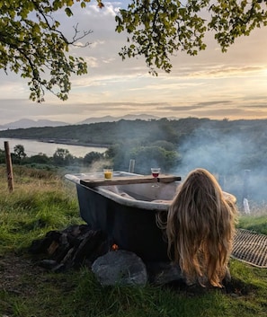 A rustic outdoor setting with a stone bathtub, a cup of tea, and a glass of juice on a table. - The Croft Collective (Oban)