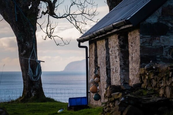 A stone building with a metal roof, a tree with a hammock, and a body of water in the background.