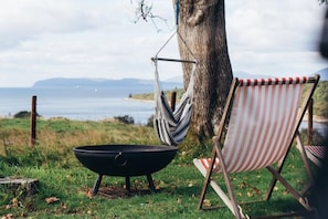 A hammock, a grill, and a striped beach chair by a tree overlooking a body of water.