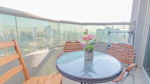 A balcony with a glass railing, a round table with chairs, and a potted plant with flowers. - The Panorama & Live Above the Creek (Ajman)