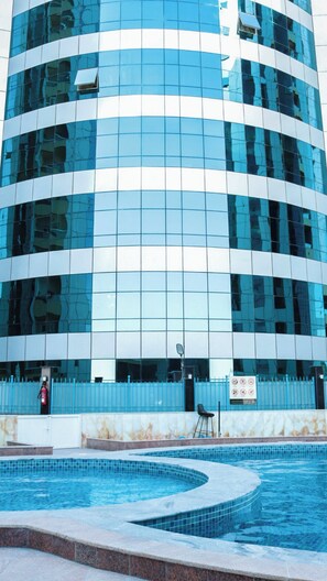 A modern building with a reflective glass facade and a swimming pool in front. - The Panorama & Live Above the Creek (Ajman)