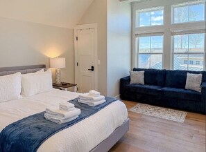 A bedroom with a bed, a nightstand, a lamp, a window, and a dark-colored sofa. - Lakeside 2-bedroom condo in awesome Laconia (Laconia)