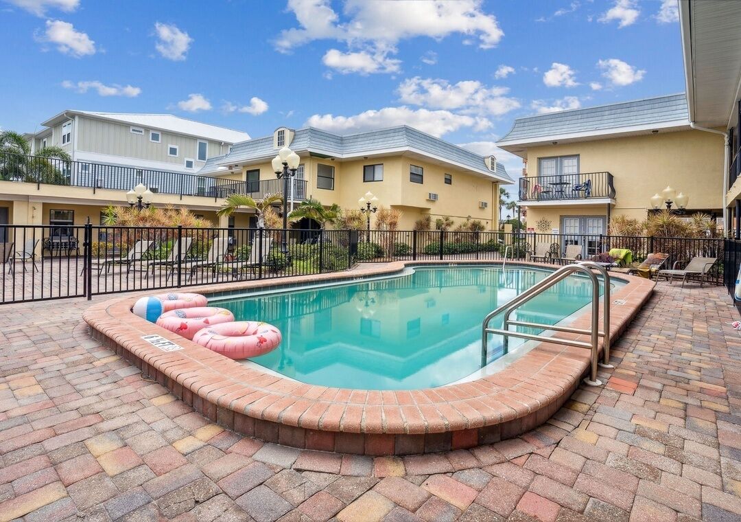 A swimming pool with a brick border, surrounded by a fence, and adjacent to a building with balconies.