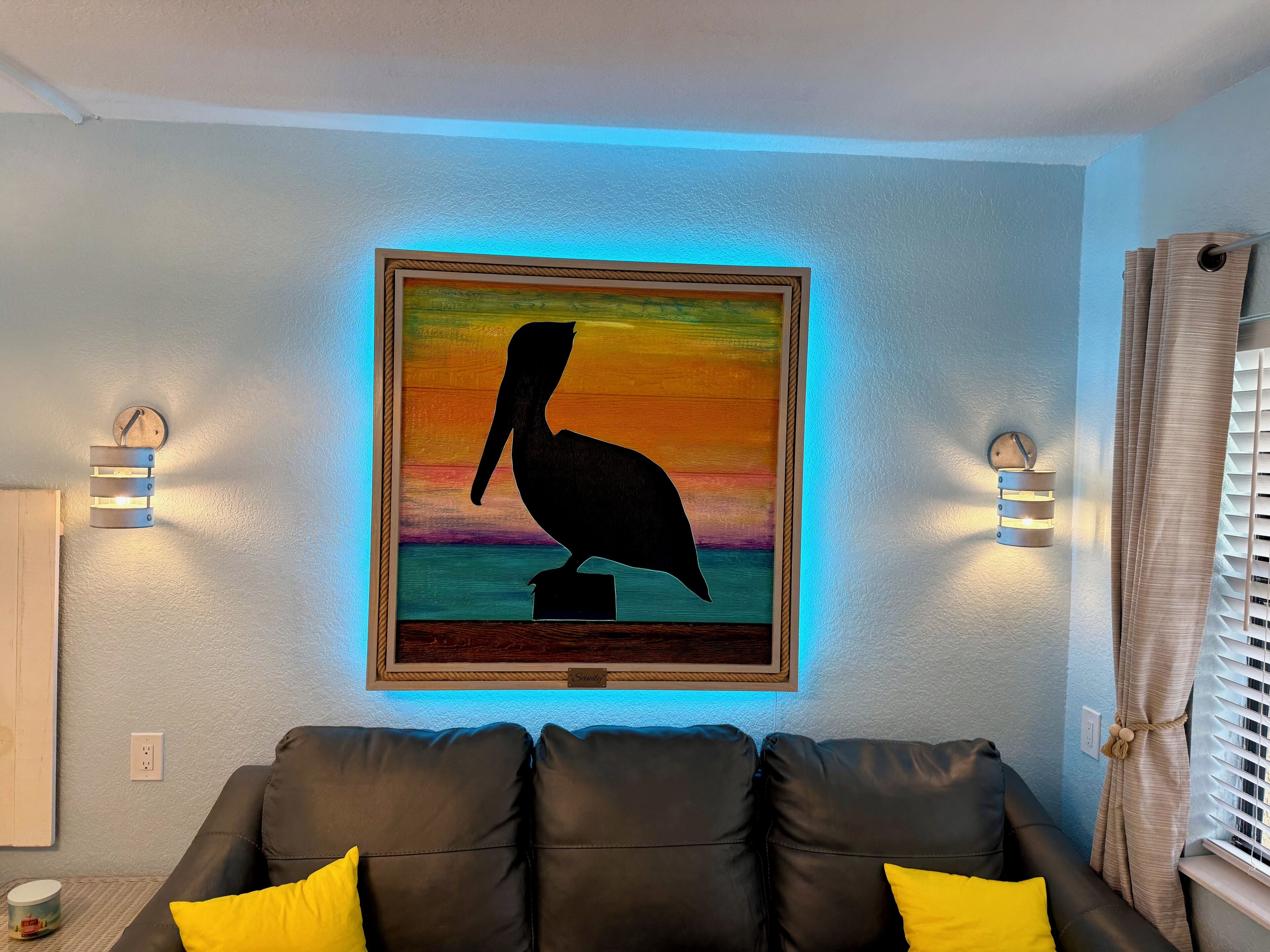 A room with a black leather sofa, a framed painting of a pelican, and two wall-mounted light fixtures.