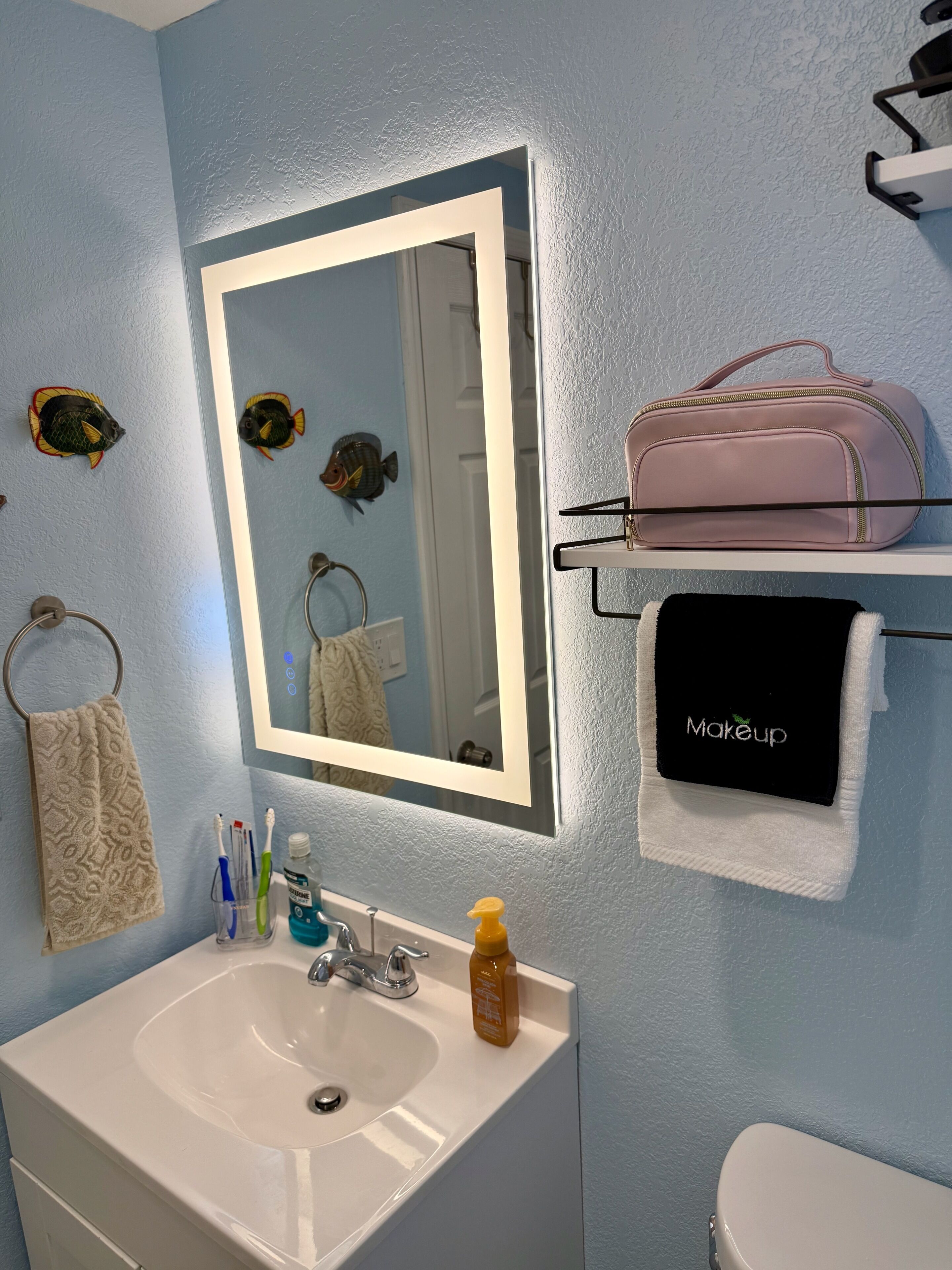 Une salle de bain avec un lavabo blanc, un miroir, un porte-serviettes et une étagère sur laquelle se trouvent un sac rose et une serviette noire avec l’inscription « Maquillage ».