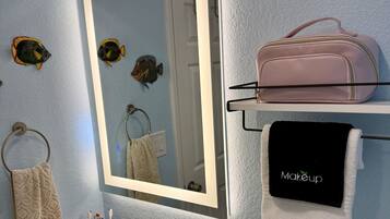 Une salle de bain avec un lavabo blanc, un miroir, un porte-serviettes et une étagère sur laquelle se trouvent un sac rose et une serviette noire avec l’inscription « Maquillage ».