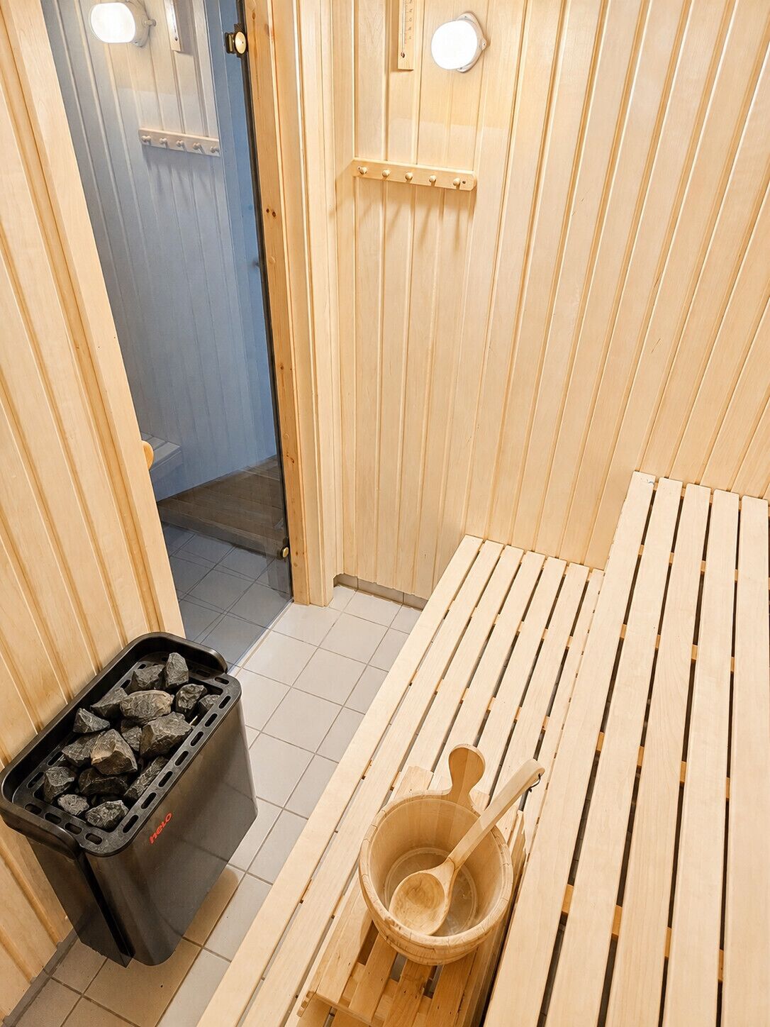 Eine Sauna mit einer Holzbank, einem Eimer und einem Holzlöffel sowie einem schwarzen Aschenbecher mit Steinen.