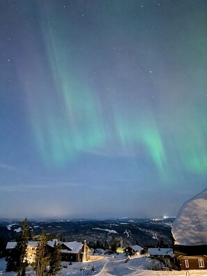 Schneebedeckte Landschaft mit einem Haus und einem großen Schneehügel, unter einem Nachthimmel mit sichtbaren Sternen und dem Polarlicht.