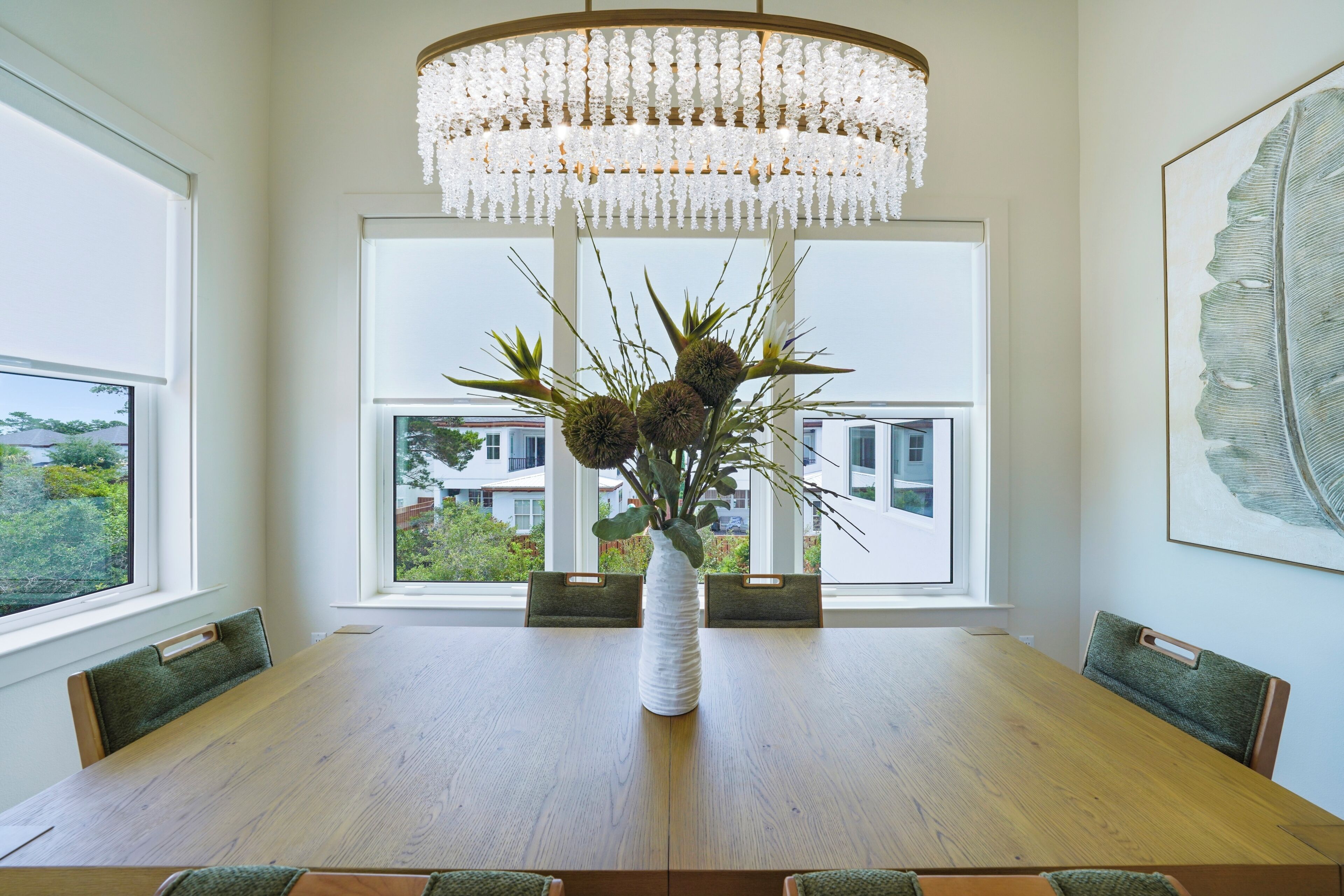 A dining area with a wooden table, a white vase with plants, and a large window with a view of buildings and greenery.