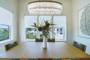A dining area with a wooden table, a white vase with plants, and a large window with a view of buildings and greenery.