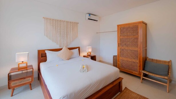 A bedroom with a wooden bed, bedside tables with lamps, a woven chair, and a wall-mounted air conditioner. -  Villa Merman | 3BR w BBQ Deck & Pool In Pererenan (Bali)