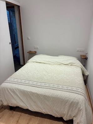 A bedroom with a bed, a wooden floor, a door leading to another room, and a small bedside table with a lamp.