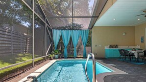 A swimming pool with a metal safety rail, a covered patio area with a table and chairs, and a view of a backyard with a fence and trees. - NEW! Beautiful home w/ private pool and golf cart! (Destin)