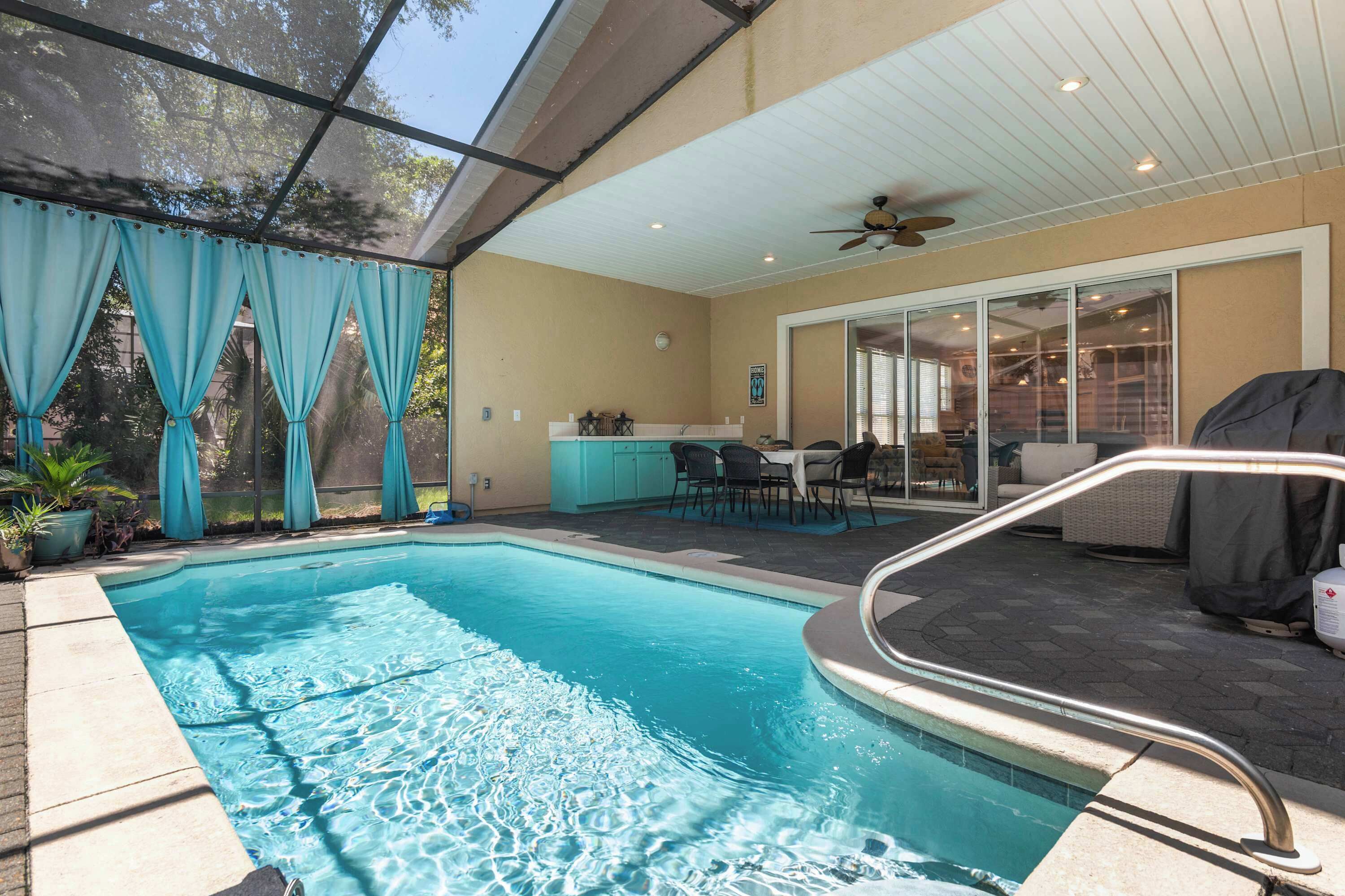 An outdoor swimming pool with a glass enclosure, a ceiling fan, and a patio area with furniture.