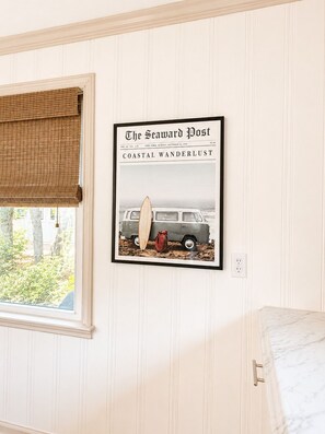 A framed newspaper article titled 'The Seaward Post' with a vintage van and a surfboard on the cover, mounted on a wall with a window with a bamboo blind. - Charming OKI Cottage Walking Distance to Beach! (Oak Island)