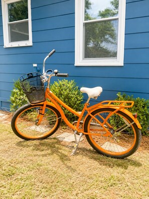 A stationary orange bicycle with a basket and a white seat, parked in front of a blue house with two windows. - Charming OKI Cottage Walking Distance to Beach! (Oak Island)