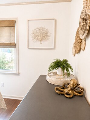 A room with a dark grey table, a framed tree artwork, a woven wall hanging, and a window with a beige blind. - Charming OKI Cottage Walking Distance to Beach! (Oak Island)