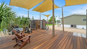 Um deck de madeira com área de estar externa, uma construção pequena e um edifício ao fundo.