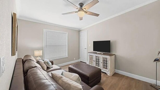 Sala de estar com sofá, um banquinho, rack para TV, ventilador de teto e cortinas.