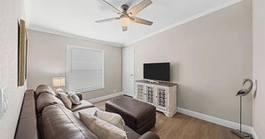 Een woonkamer met een bank, voetenbankje, tv-meubel, plafondventilator en jaloezieën.