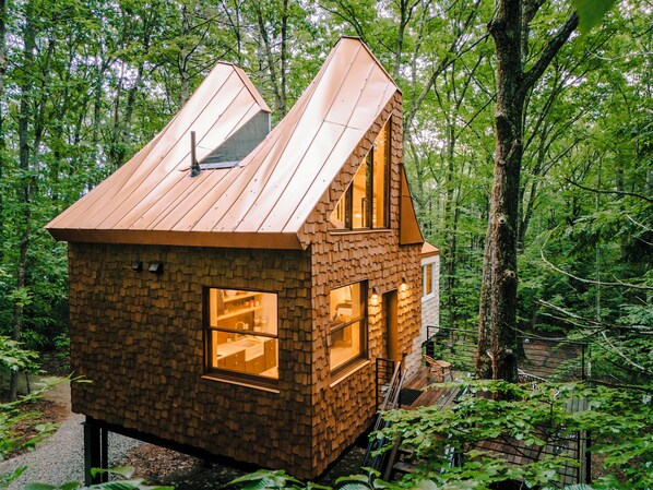 Una pequeña cabaña de madera con techo inclinado, ventanas grandes y una chimenea, rodeada por un bosque.