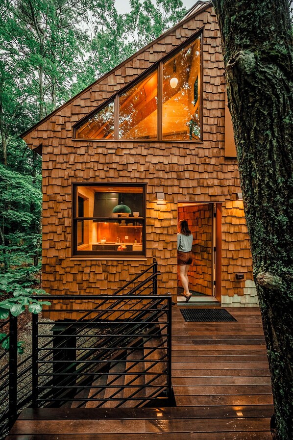 A wooden cabin with a large window, a staircase, and a view of the surrounding forest.
