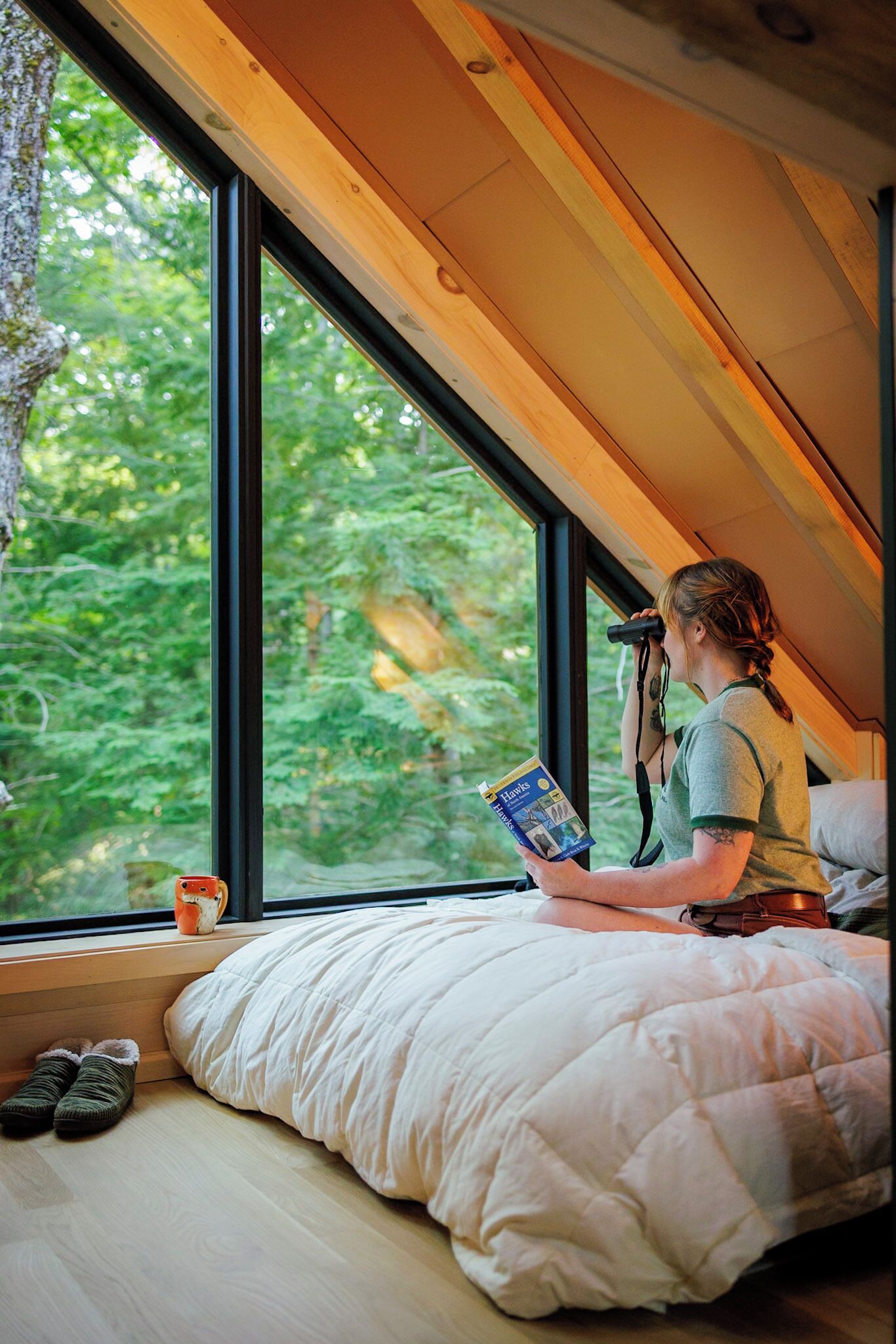Eine Person sitzt auf einem Bett in einem Zimmer mit einem großen Fenster und liest ein Buch.