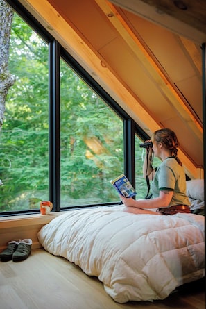 Una persona sentada en una cama en una habitación con un ventanal grande, leyendo un libro.