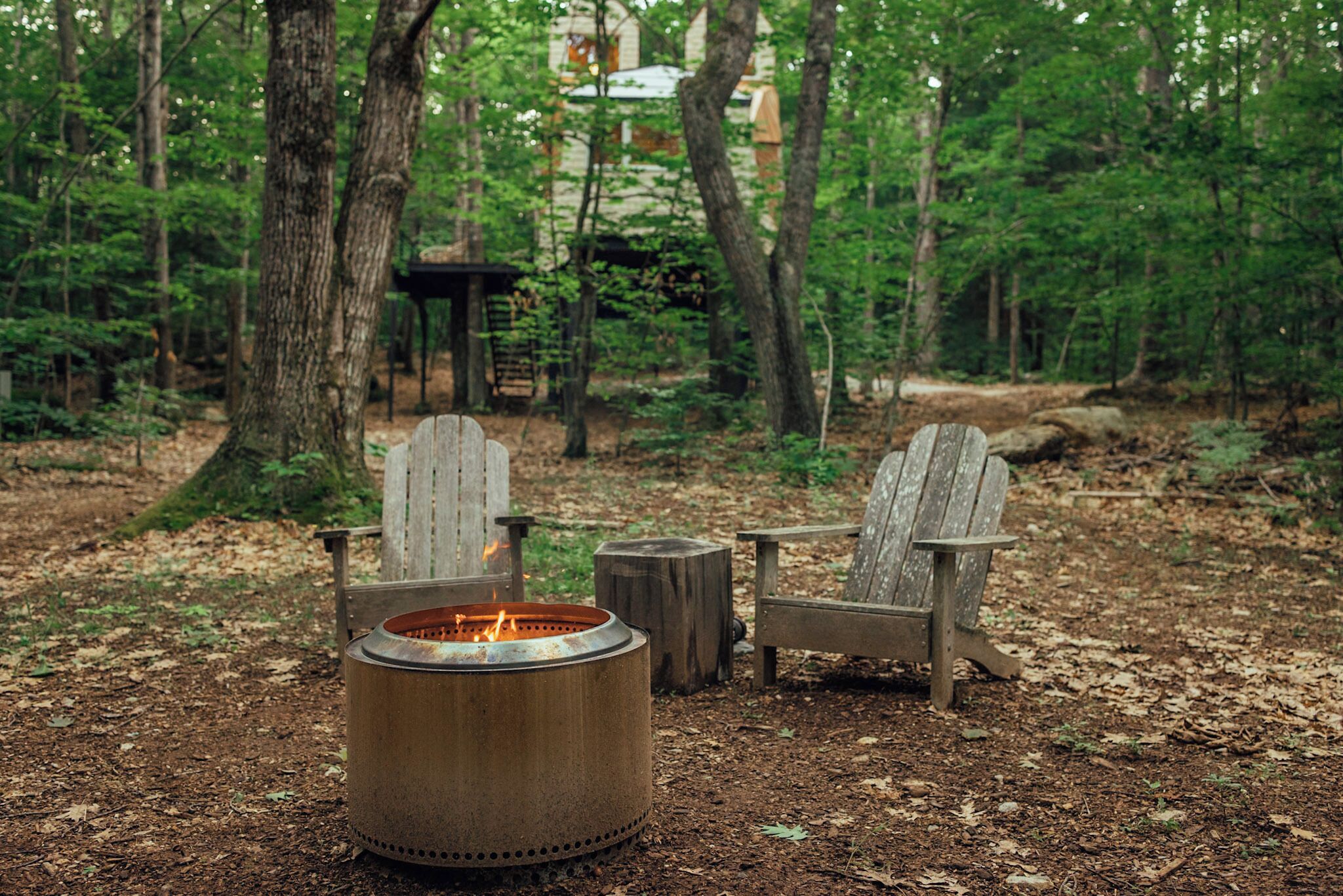 Waldszene mit einem Feuerkorb aus Metall und zwei Adirondack-Stühlen.