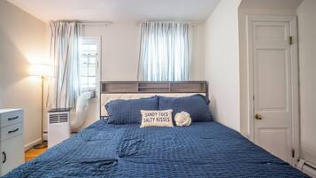 A bedroom with a bed, a bedside table, a lamp, a window with curtains, and a door.