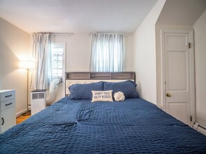 A bedroom with a bed, a bedside table, a lamp, a window with curtains, and a door. - Connecticut Beach Home (Old Lyme)