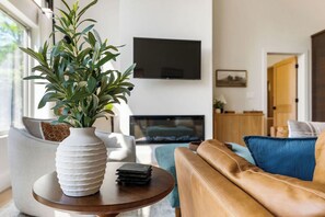 A living room with a sofa, a round wooden table with a white vase and a book, a television mounted on the wall, and a door leading to another room. - !New!-Sunset Escape- Bayside Bluff with Hot Tub (Egg Harbor)