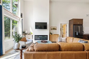 A living room with a large window, a flat-screen TV, a sofa, and a kitchen area in the background. - !New!-Sunset Escape- Bayside Bluff with Hot Tub (Egg Harbor)