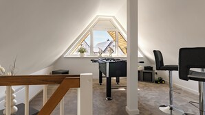 A modern loft bedroom with a sloped ceiling, a foosball table, a black office chair, and a window overlooking a neighboring building. - A Getaway in Büsum - Vacation Home with Sauna & Fireplace (Büsum)