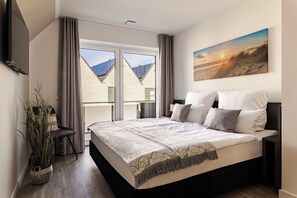 Ein modernes Schlafzimmer mit einem großen Bett, einem Flachbildfernseher, einem Bild von einem Strand und Blick auf ein Gebäude mit Solaranlage.